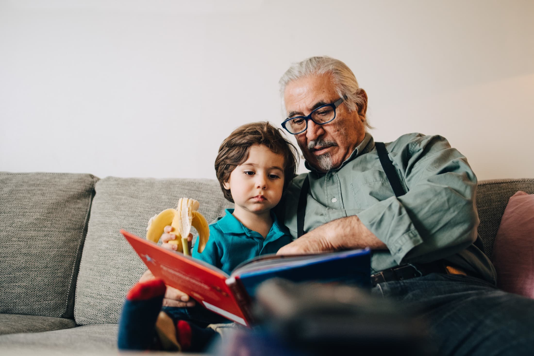 A grandfather reading a book with his grandson on the sofa at home